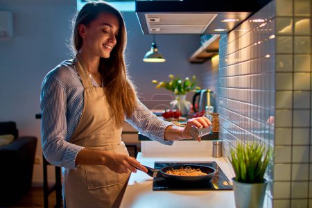 Photo for Happy smiling attractive young cooking woman housewife salting and preparing food in a frying pan on the stove for evening dinner at modern loft style kitchen - Royalty Free Image
