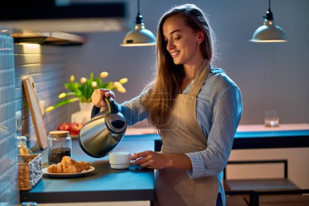 Téléchargez les photos : Joyeux sourire attrayant jeune femme au foyer verser et faire du thé à l'aide d'une bouilloire électrique pour la pause café du soir à la cuisine moderne de style loft - en image libre de droit