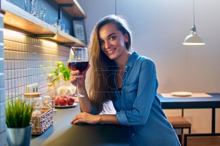 Foto de Retrato de hermosa sonriente linda bebedora sommelier sosteniendo una copa de vino tinto y sentado en la moderna cocina loft en casa - Imagen libre de derechos