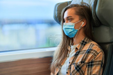 Photo for Young woman passenger in a medical mask and white headphones while safety traveling by train during coronavirus epidemic. Virus protection and wearing mask in public transport - Royalty Free Image