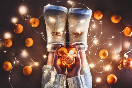 Photo for Ripe mandarins orange in hands, warm white christmas lights garland and female legs wearing warm fluffy soft winter slippers at cozy home at christmas eve. Top view - Royalty Free Image