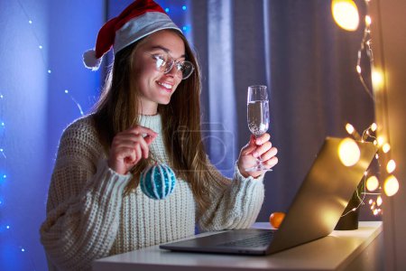 Foto de Joven feliz sonriente alegre mujer de Navidad con sombrero de santa beber champán y hablar en línea en videollamada virtual mientras se celebra la fiesta de Navidad remota en un ordenador portátil en casa - Imagen libre de derechos