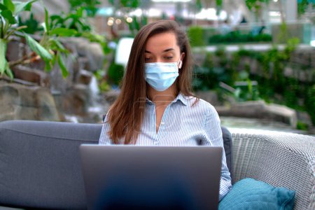 Foto de Moderna mujer freelancer inteligente que usa mascarilla médica trabajando remotamente en línea en una computadora en un lugar público - Imagen libre de derechos