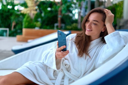 Foto de Mujer sonriente atractiva joven que usa albornoz blanco usando un teléfono inteligente para ver video y navegar en línea mientras está acostada en una tumbona durante la relajación en el balneario - Imagen libre de derechos