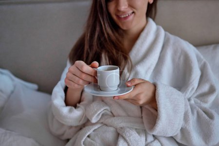 Foto de Mujer perezosa en albornoz acostado en la cama y disfrutar de un buen desayuno por la mañana con taza de café aromático durante la relajación en el acogedor dormitorio confort en la habitación del hotel. Un estilo de vida fácil. Comienzo del nuevo día - Imagen libre de derechos