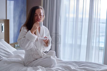 Foto de Mujer perezosa en albornoz acostado en la cama y disfrutar de un buen desayuno por la mañana con taza de café aromático durante la relajación en el acogedor dormitorio confort en la habitación del hotel. Un estilo de vida fácil. Comienzo del nuevo día - Imagen libre de derechos