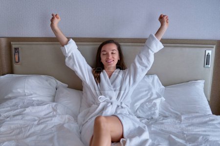 Téléchargez les photos : Happy souriant femme paresseuse joyeuse avec les mains levées en peignoir se réveiller d'un bon sommeil le matin lors de la détente dans la chambre confortable confort à la chambre d'hôtel. Style de vie facile - en image libre de droit