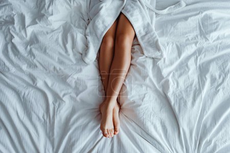 Téléchargez les photos : Femme paresseuse portant un peignoir avec de longues jambes fines et douces couchées sur un lit blanc et se relaxant dans une chambre confortable de confort à la chambre d'hôtel. Style de vie facile - en image libre de droit