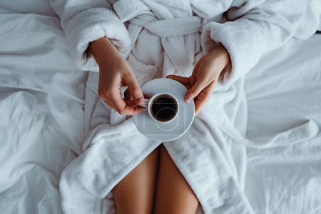 Foto de Mujer perezosa con albornoz acostado en la cama y disfrutando de la taza de café aromático durante la relajación en el acogedor dormitorio confort en la habitación del hotel. Un estilo de vida fácil. Comienzo del nuevo día - Imagen libre de derechos
