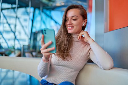 Photo for Young modern casual happy joyful woman wearing wireless headphones using smartphone for watching video and reading news online while sitting and resting at cafe - Royalty Free Image