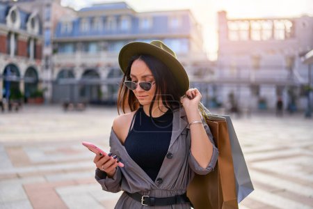 Téléchargez les photos : Élégant mode jeune femme hipster shopaholic portant un chapeau en feutre et des lunettes de soleil avec des sacs à provisions en papier en utilisant le téléphone et des promenades dans le centre d'une ville européenne - en image libre de droit