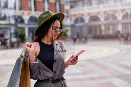Téléchargez les photos : Élégant mode jeune femme hipster shopaholic portant un chapeau en feutre et des lunettes de soleil avec des sacs à provisions en papier en utilisant le téléphone et des promenades dans le centre d'une ville européenne - en image libre de droit