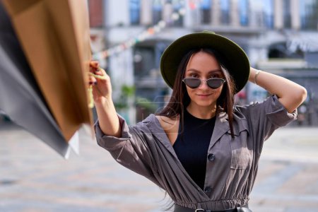 Foto de Retrato de mujer hipster elegante y sonriente joven shopaholic usando sombrero de fieltro y gafas de sol con bolsas de compras de papel durante descuentos de venta de viernes negro - Imagen libre de derechos