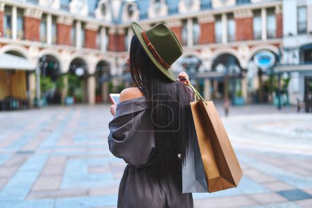 Téléchargez les photos : Élégant à la mode hipster brunette femme shopaholic avec des cheveux longs chapeau en feutre avec des sacs à provisions en papier promenades dans le centre d'une ville européenne pendant la semaine de vente du vendredi noir - en image libre de droit