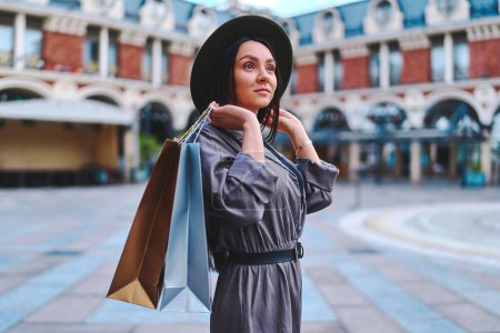 Téléchargez les photos : Élégant à la mode jeune mignon attrayant hipster femme shopaholic porter chapeau en feutre avec des sacs à provisions en papier promenades dans le centre d'une ville européenne pendant la semaine de vente du vendredi noir - en image libre de droit