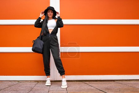 Foto de Elegante mujer deportiva joven y moderna con mono negro, sombrero de panama, top blanco corto y botas sosteniendo una bolsa de gimnasio y de pie contra una pared naranja brillante - Imagen libre de derechos