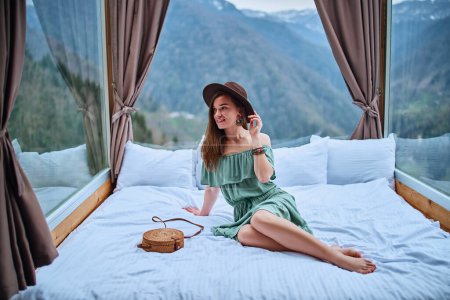 Photo for Portrait of young charming smiling beautiful boho chic brunette woman traveler with long legs wearing bare shoulder emerald dress and felt brown hat sitting on bed with mountain view - Royalty Free Image