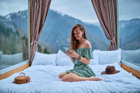Photo for Young charming smiling beautiful boho chic brunette woman traveler wearing bare shoulder emerald dress sitting on bed at hotel room with mountain view and using phone for online communication - Royalty Free Image