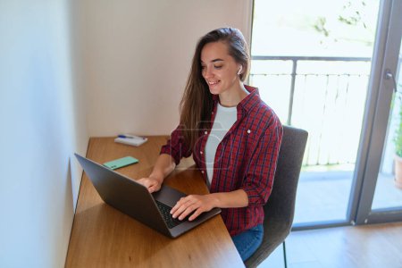 Photo for Happy young casual smart modern smiling girl freelancer with wireless headphone using laptop during remote working online at home - Royalty Free Image
