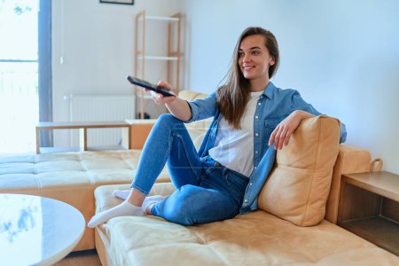 Photo for Smiling casual modern joyful happy young girl sitting on a sofa, holding remote and watching TV a t home - Royalty Free Image
