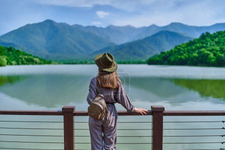 Foto de Chica viajera de pie solo en el borde del muelle y mirando al lago y las montañas. Disfrutando de feliz momento de libertad la vida y sereno ambiente tranquilo y tranquilo en la naturaleza. Vista trasera - Imagen libre de derechos