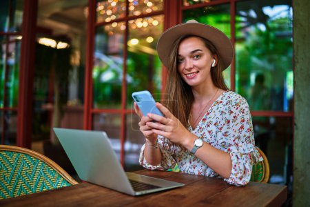 Photo for Happy cheerful smiling beautiful cute joyful young freelancer girl wearing wireless headphones using laptop and phone for remote work and online browsing at green cafe - Royalty Free Image