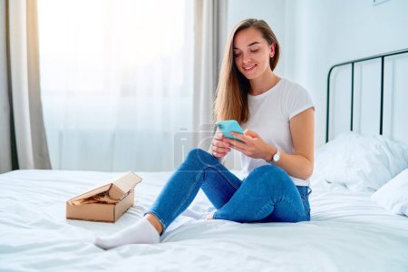 Photo for Happy joyful young satisfied shopaholic woman client sitting on a bed at home with received carton parcel box after online ordering, easy and fast service commerce delivery concept - Royalty Free Image