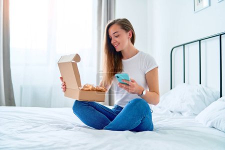 Photo for Happy joyful young satisfied shopaholic woman client sitting on a bed at home and opening received carton parcel box after online ordering, easy and fast service commerce delivery concept - Royalty Free Image
