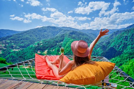 Photo for Young carefree girl traveler wearing hat relaxing with a glass of wine and lying on a hammock during enjoying free happy moment life against the background of green big mountains - Royalty Free Image
