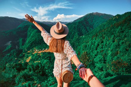 Photo for Follow me concept and traveling together. Girl traveler wearing hat, round straw bag and short jumpsuit holds the boyfriend's hand and leading to green big mountains vacation - Royalty Free Image