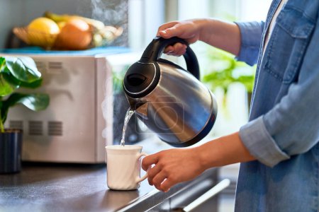 Téléchargez les photos : Mains féminines versant l'eau bouillante d'une bouilloire moderne en métal inoxydable dans une tasse en verre pour infuser le thé dans la cuisine à la maison - en image libre de droit