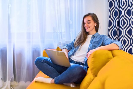 Photo for Young attractive smiling happy cute millennial girl sitting on the couch using a laptop and wireless headphones for watching video webinar online, video call and learning language courses at home - Royalty Free Image