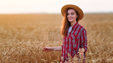 Foto de Retrato de linda joven feliz mujer sonriente agricultora de pie sola durante el caminar a través de un campo amarillo de trigo seco maduro entre espiguillas doradas al atardecer - Imagen libre de derechos