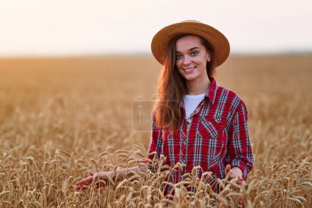 Foto de Retrato de linda joven satisfecha feliz sonriente hermosa agricultora de pie sola durante el caminar a través de un campo amarillo de trigo seco maduro entre espiguillas de oro al atardecer - Imagen libre de derechos