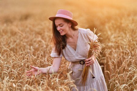 Photo for Happy smiling attractive cute free young female wearing hat and dress standing in golden yellow wheat field and enjoying serene beautiful moment life at summertime - Royalty Free Image