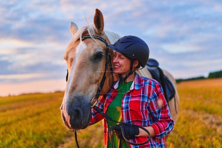 Photo for Young cute happy joyful satisfied smiling woman rider wearing helmet hugging and stroking beautiful blonde palomino horse at meadow at sunset at golden hour time - Royalty Free Image