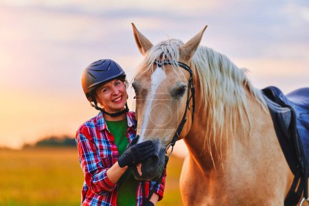 Foto de Joven linda feliz alegre satisfecha sonriente jinete con casco abrazando y acariciando hermoso caballo palomino rubia en el prado al atardecer a la hora dorada - Imagen libre de derechos