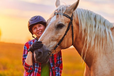 Foto de Joven linda feliz alegre satisfecha sonriente jinete con casco abrazando y acariciando hermoso caballo palomino rubia en el prado al atardecer a la hora dorada - Imagen libre de derechos