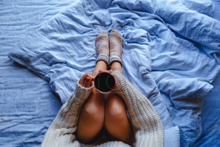 Téléchargez les photos : Femme confortable dans un pull chaud et chaussettes tricotées assis sur un lit blanc doux et tenant une tasse de chocolat chaud dans ses mains. Vue du dessus - en image libre de droit