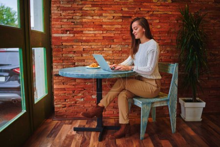 Foto de Lindo joven inteligente copywriter moderno independiente chica tipos en un teclado portátil durante el trabajo en línea de forma remota en un café - Imagen libre de derechos