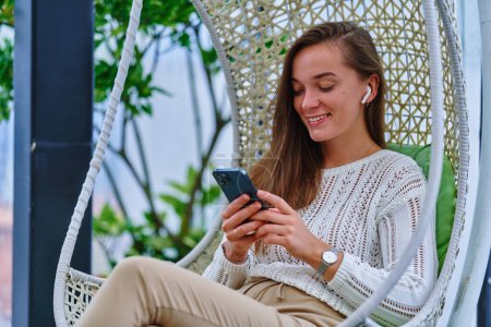 Foto de Linda joven feliz alegre sonriente mujer moderna inteligente con auriculares blancos usando el teléfono móvil para navegar, chat en línea y redes sociales - Imagen libre de derechos