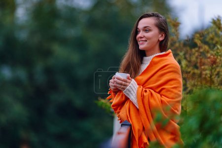 Foto de Hermosa linda feliz sola sonriente mujer envuelta en una manta a cuadros de pie en la terraza y sosteniendo una taza de bebida caliente durante el tiempo de descanso cómodo y acogedor - Imagen libre de derechos