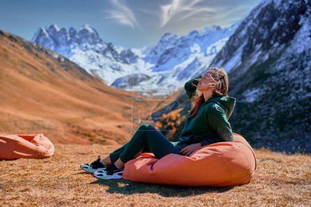 Foto de Chica vagabunda sentada en una silla suave durante el descanso en un camping. Viajero disfrutando de paisaje paisajístico mientras viaja en un valle de montaña - Imagen libre de derechos