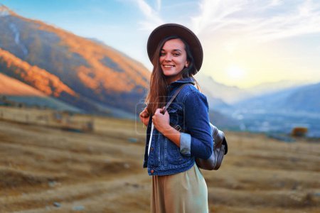 Téléchargez les photos : Portrait de heureux joyeux jeune fille hipster sourire mignon vagabond portant chapeau et sac à dos voyageant seul dans la vallée de la montagne en Géorgie pays - en image libre de droit