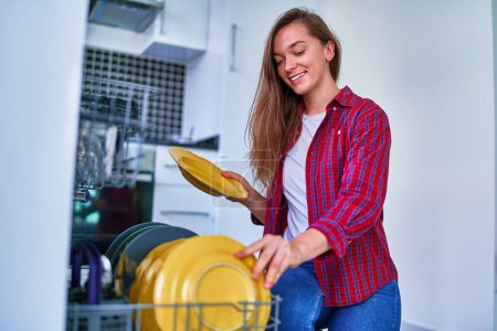Photo for Young joyful happy satisfied smiling cute housewife woman using modern dishwasher for wash dishes and glasses at white home kitchen - Royalty Free Image