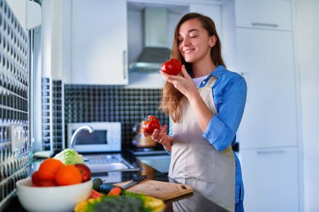 Photo for Young happy joyful cute smiling housewife wearing apron preparing fresh vegetarian salad for healthy eating at home kitchen - Royalty Free Image