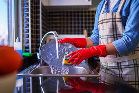 Foto de Ama de casa con delantal y guantes rojos de goma protectora lavar los platos en casa en la cocina - Imagen libre de derechos