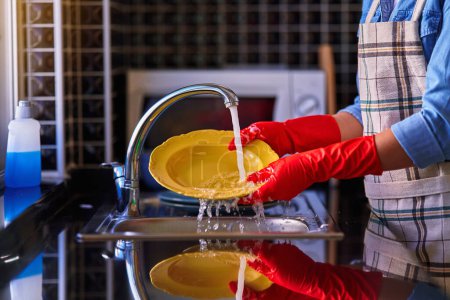 Téléchargez les photos : Femme au foyer portant un tablier et des gants en caoutchouc rouge pour laver la vaisselle à la maison dans la cuisine - en image libre de droit