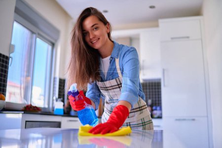 Photo for Happy cute caucasian attractive young joyful smiling housewife woman wipes and disinfects dust table using spray bottle and rag - Royalty Free Image