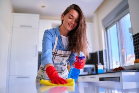 Téléchargez les photos : Joyeux mignon caucasien attrayant jeune femme au foyer souriante joyeuse essuie et désinfecte la table de poussière à l'aide de vaporisateur et chiffon - en image libre de droit